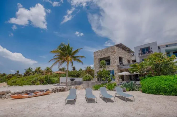 Four sun loungers on a sandy tropical beach in front of a stone beachfront villa with a palm tree and kayak under a bright blue sky