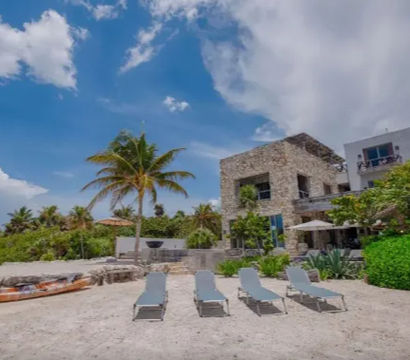 Four sun loungers on a sandy tropical beach in front of a stone beachfront villa with a palm tree and kayak under a bright blue sky