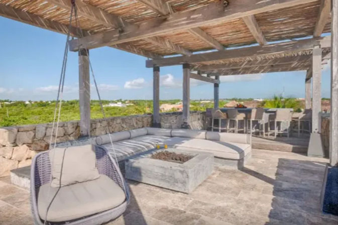 Rooftop pergola patio with hanging wicker chair, cushioned L-shaped outdoor sofa around a concrete fire pit, stone tile floor and bar seating, overlooking green coastal landscape and blue sky
