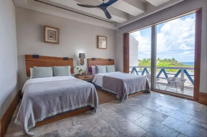 Coastal bedroom with two double beds, wooden headboards and gray linens, sliding glass doors to a sunny balcony overlooking tropical greenery and the blue ocean.