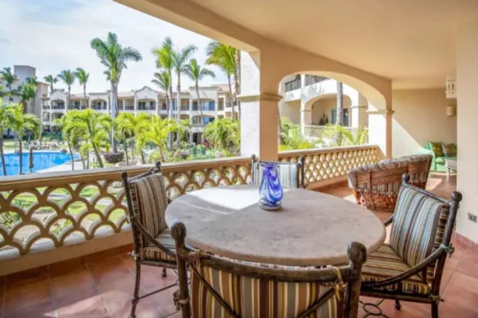Sunlit tropical resort balcony with round stone table, striped cushioned chairs and blue glass vase, overlooking palm trees, swimming pool and stucco guest buildings.
