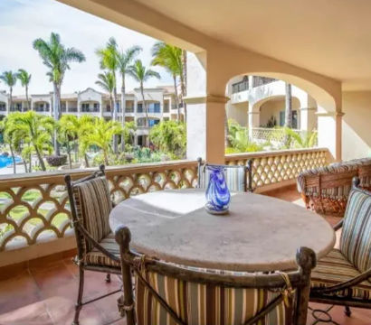 Sunlit tropical resort balcony with round stone table, striped cushioned chairs and blue glass vase, overlooking palm trees, swimming pool and stucco guest buildings.