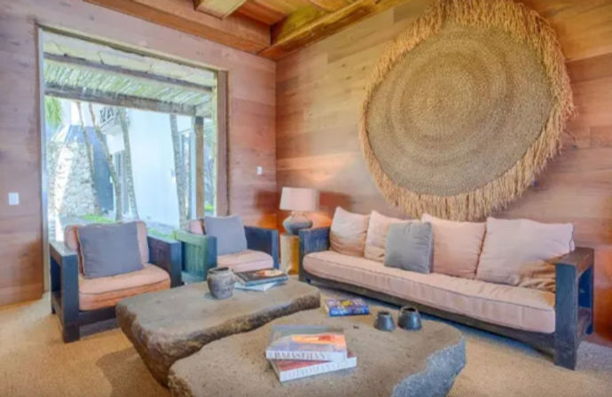 Cozy tropical living room with wood-paneled walls, oversized round woven wall art, beige sofa and two cushioned armchairs, rough stone coffee tables with books and pottery, and an open doorway framing palm trees outside.