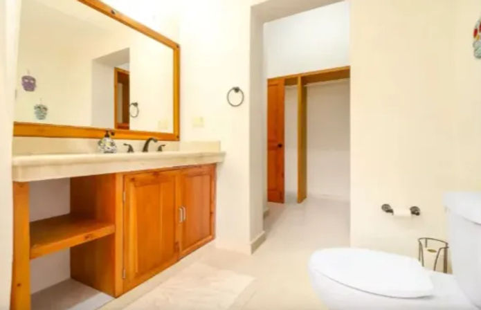 Bright residential bathroom with a wooden vanity and large framed mirror, cream countertop, open doorway to a walk-in closet, white tile floor and toilet.