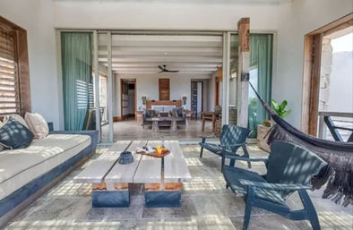 Sunlit coastal villa patio with cushioned daybed, rustic wooden coffee table, two blue lounge chairs and a hammock opening through sliding glass doors into a bright living room — inviting outdoor lounge space.