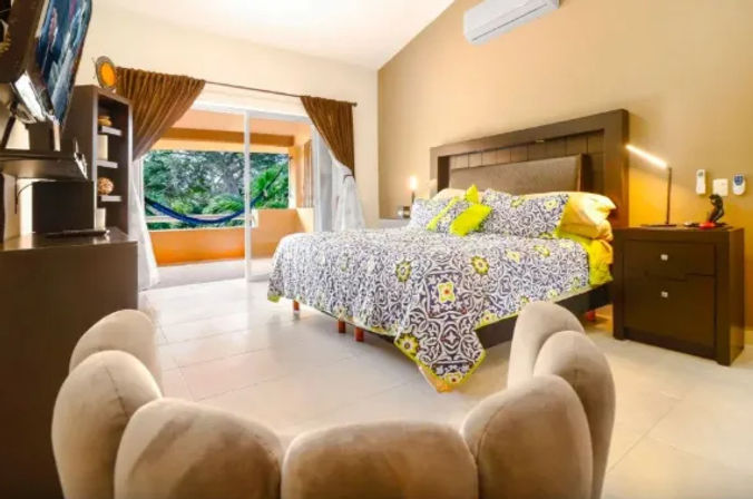 Sunlit tropical bedroom with king bed in blue-and-yellow patterned linens, dark wood nightstands and TV, and sliding glass doors opening to a balcony with a hammock and lush greenery.