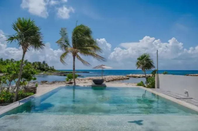Oceanfront infinity pool overlooking a turquoise tropical sea, framed by swaying palm trees, a beach umbrella on the sand and a bright blue sky with fluffy clouds.