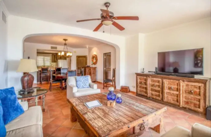 Bright Mediterranean-style living room with terracotta tile floors, rustic wooden coffee table and TV console, beige sofas with blue accent pillows, ceiling fan, and an arched open-concept dining area visible in the background.