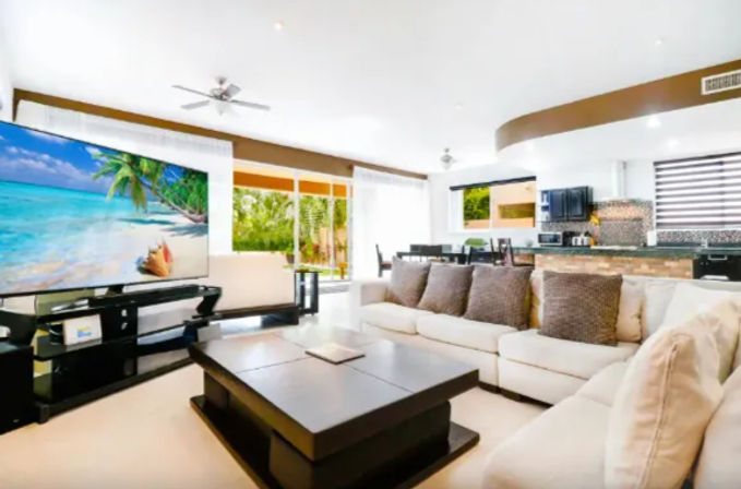 Bright open-plan living room and kitchen with a large flat-screen TV showing a beach, beige sectional sofa with brown throw pillows, dark wood coffee table, dining area, ceiling fans, and sliding glass doors opening to a lush tropical garden.