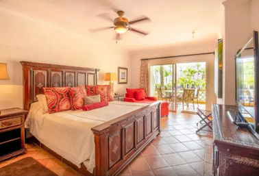 Sunlit tropical resort-style king bedroom with carved wood bed, red patterned pillows, ceiling fan and sliding balcony doors opening to a palm-tree courtyard.