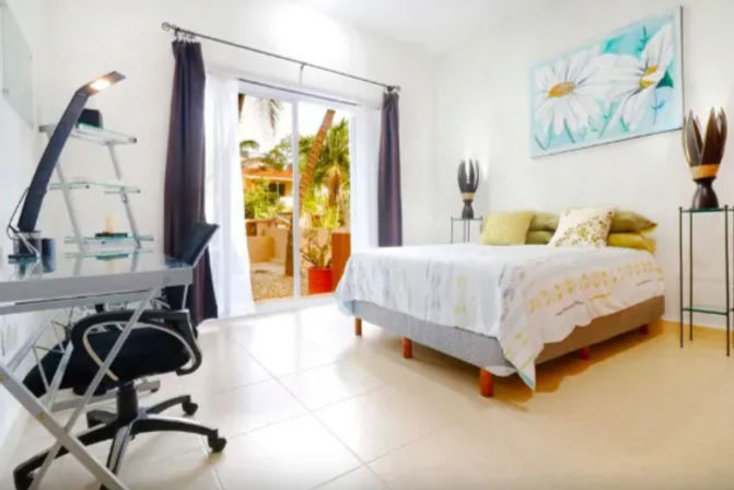 Sunny modern bedroom with a cozy bed, work-from-home glass desk and chair, decorative lamps and sliding glass doors opening to a tropical patio with palm trees.