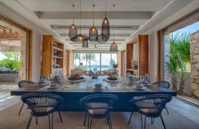 Open-plan beachfront dining room with a long wooden table set for a meal, black woven chairs, sculptural pendant lights, sliding walls framing palm trees and turquoise ocean views — tropical coastal villa vibe.