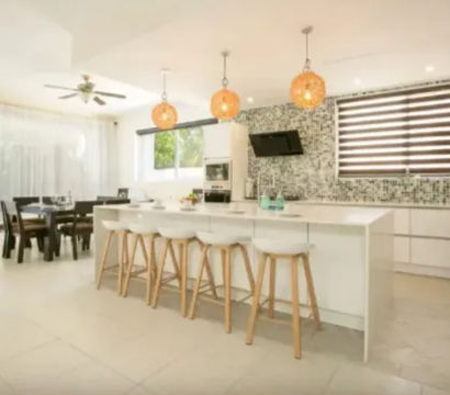 Bright, sunlit suburban open-plan kitchen and dining area with a long white island and four wooden bar stools, three woven pendant lights, mosaic tile backsplash, stainless appliances, dark-wood dining table, ceiling fan, and sliding glass doors to a green lawn.