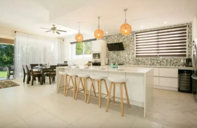 Bright, sunlit suburban open-plan kitchen and dining area with a long white island and four wooden bar stools, three woven pendant lights, mosaic tile backsplash, stainless appliances, dark-wood dining table, ceiling fan, and sliding glass doors to a green lawn.