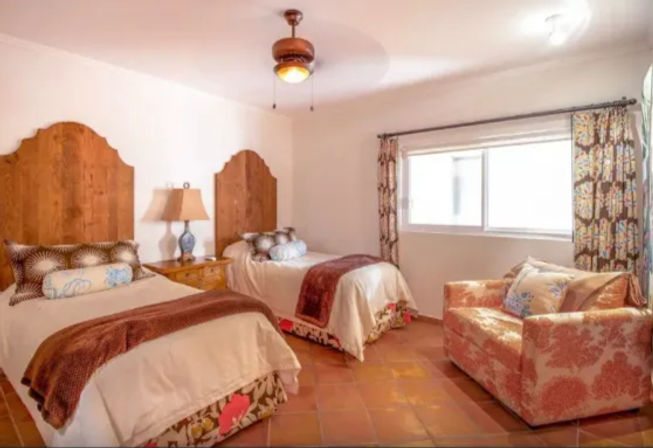 Cozy sunlit guest bedroom with two twin beds, carved wooden headboards, terracotta tile floor, patterned curtains, bedside lamp and a floral armchair.