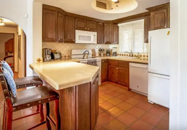 Cozy kitchen with warm wood cabinets, cream countertop breakfast bar with two leather barstools, white appliances (fridge, microwave, dishwasher), terracotta tile floor and window blinds.