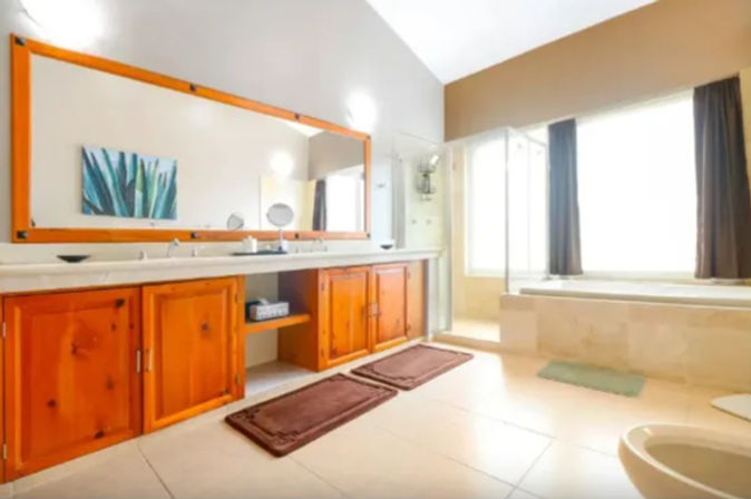 Bright, spacious master bathroom with double-sink vanity, warm wood cabinets, large framed mirror, glass walk-in shower and sunlit bathtub by a window