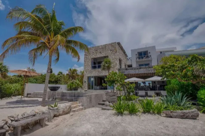 Tropical beachfront stone villa with a palm tree on sandy shore, ocean-view patio, outdoor dining umbrellas and lush greenery under a bright blue sky