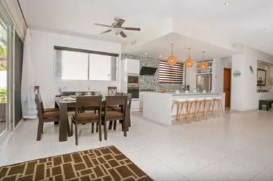 Modern open-plan kitchen and dining room with white tile floors, a large marble island with wooden bar stools and pendant lights, dark wood dining table, ceiling fan and sliding glass doors to the patio.