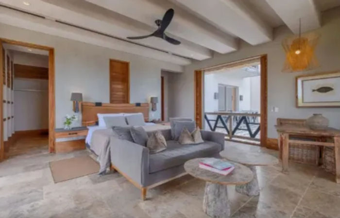 Relaxing coastal bedroom suite with king bed, gray loveseat, stone tile floor, wooden accents, ceiling fan and private balcony terrace