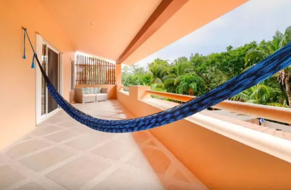 Covered tropical balcony with bright orange stucco walls, a blue hammock stretched across the terrace, cushioned seating and lush palm-tree view