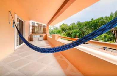 Covered tropical balcony with bright orange stucco walls, a blue hammock stretched across the terrace, cushioned seating and lush palm-tree view