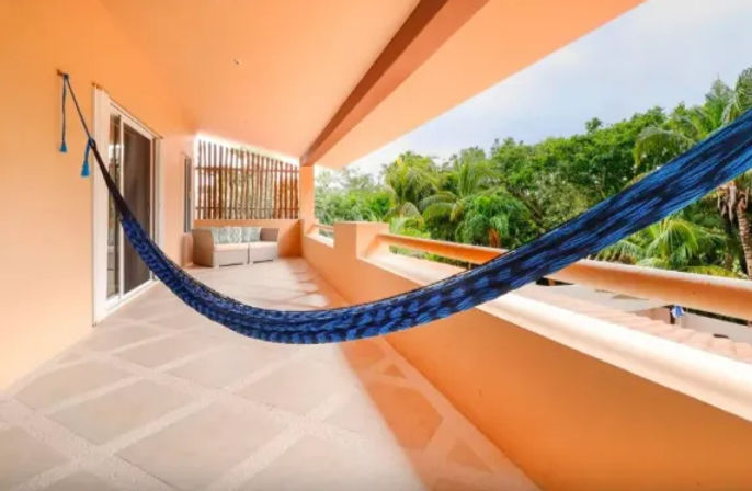 Covered tropical balcony with bright orange stucco walls, a blue hammock stretched across the terrace, cushioned seating and lush palm-tree view