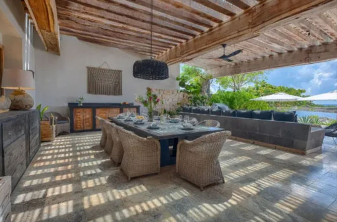 Covered coastal patio dining area with long table set for a meal, woven rattan chairs, exposed wooden beam ceiling, stone tile floor and open view of tropical greenery and ocean umbrellas.
