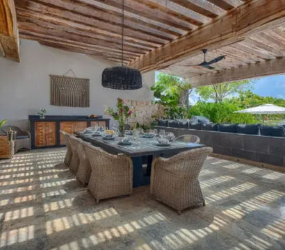 Covered coastal patio dining area with long table set for a meal, woven rattan chairs, exposed wooden beam ceiling, stone tile floor and open view of tropical greenery and ocean umbrellas.