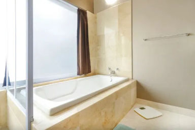 Spa-like beige-tiled bathroom with a built-in white soaking tub beneath a frosted window and brown curtain, glass shower screen, wall-mounted taps, towel rail and a digital floor scale.