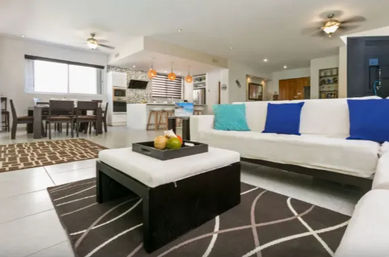 Modern open-plan living room and kitchen with white sofas, blue and turquoise throw pillows, dark ottoman with a fruit tray, patterned area rugs, dining table in the background, and pendant lights over the kitchen island.