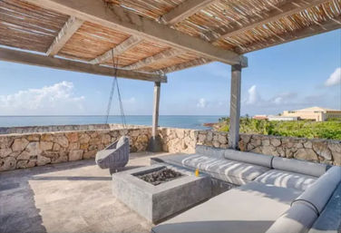 Relaxing covered seaside terrace with woven hanging chair, concrete fire pit and cushioned L-shaped lounge behind a low stone wall, overlooking turquoise ocean and rocky tropical coastline under a bright blue sky.