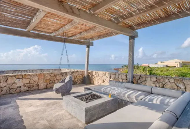 Relaxing covered seaside terrace with woven hanging chair, concrete fire pit and cushioned L-shaped lounge behind a low stone wall, overlooking turquoise ocean and rocky tropical coastline under a bright blue sky.