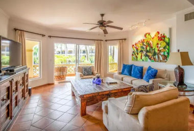 Sunny tropical condo living room with terracotta tile floors, beige sofas with blue throw pillows, rustic wooden coffee table, ceiling fan, sliding glass doors to a balcony with palm trees, and bright cactus wall art.