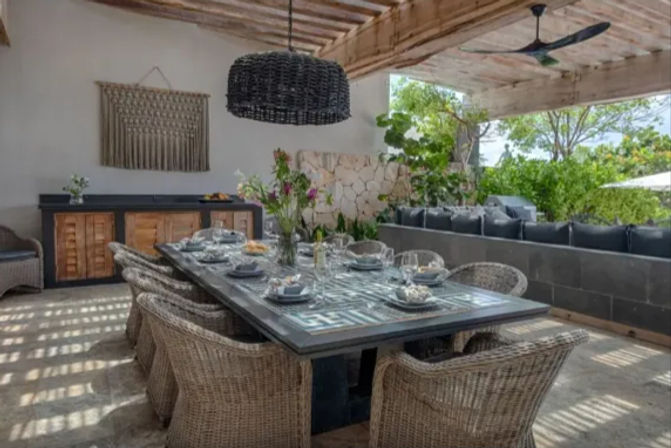Inviting covered outdoor dining patio with a long tile-top table set for eight, wicker chairs, black woven pendant light, cushioned lounge seating and lush garden views.