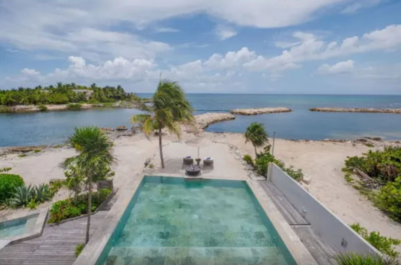 Tropical beachfront infinity pool overlooking turquoise ocean and sandy shore with swaying palm trees and rocky breakwaters under a bright blue sky.