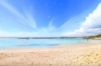 Sunny tropical beach with soft white sand, calm turquoise water and a bright blue sky streaked with wispy clouds, shallow shoreline and a distant person wading — idyllic vacation scene.