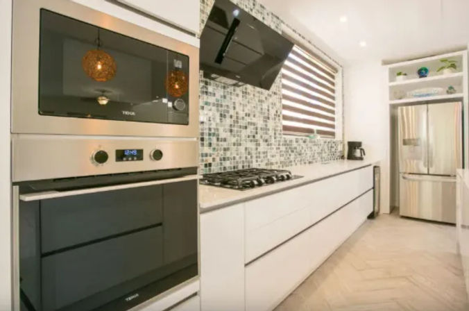 Bright modern galley kitchen with white flat-panel cabinets, stainless steel built-in oven and microwave, gas cooktop under a black hood, mosaic glass tile backsplash, striped window blind and stainless French-door refrigerator.