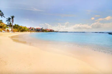 Tropical sandy beach with palm trees, turquoise calm water and resort buildings along the shoreline under a sunny blue sky