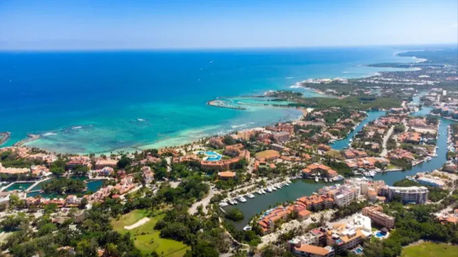 Aerial view of a sunny tropical coastal resort town with turquoise ocean, sandy beaches, winding canals and marinas dotted with boats, lush greenery and low-rise waterfront buildings.