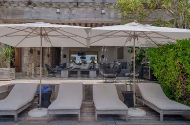Four cushioned sun loungers under white patio umbrellas on a wooden deck, facing a shaded modern outdoor living area with gray sofas, wicker chairs, blue towels and lush greenery — a relaxing coastal-style patio.