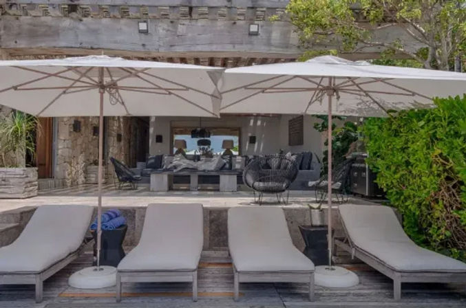Four cushioned sun loungers under white patio umbrellas on a wooden deck, facing a shaded modern outdoor living area with gray sofas, wicker chairs, blue towels and lush greenery — a relaxing coastal-style patio.