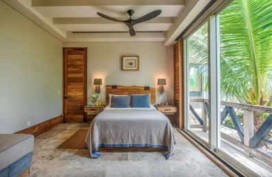 Cozy tropical beachfront bedroom with queen bed, wooden headboard, matching nightstands and lamps, ceiling fan, and large sliding glass doors opening to a palm-fringed balcony