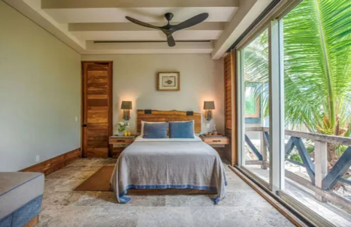 Cozy tropical beachfront bedroom with queen bed, wooden headboard, matching nightstands and lamps, ceiling fan, and large sliding glass doors opening to a palm-fringed balcony