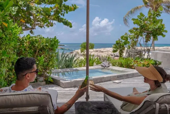 Two people relaxing on lounge chairs under an umbrella, toasting with drinks beside a private plunge pool and turquoise ocean, framed by tropical greenery and palm trees.