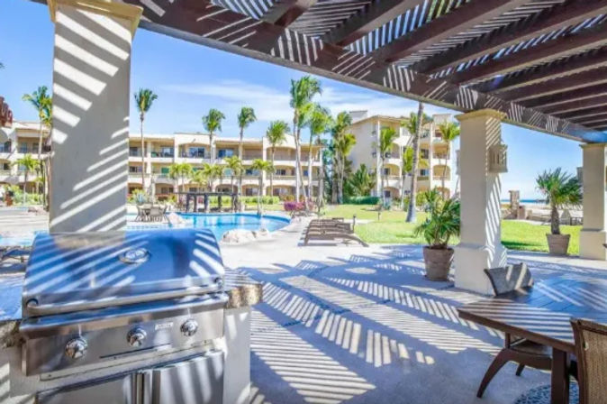 Sunlit beachfront resort patio with striped pergola shadows, stainless-steel grill, poolside lounge chairs, tropical palm trees and ocean view