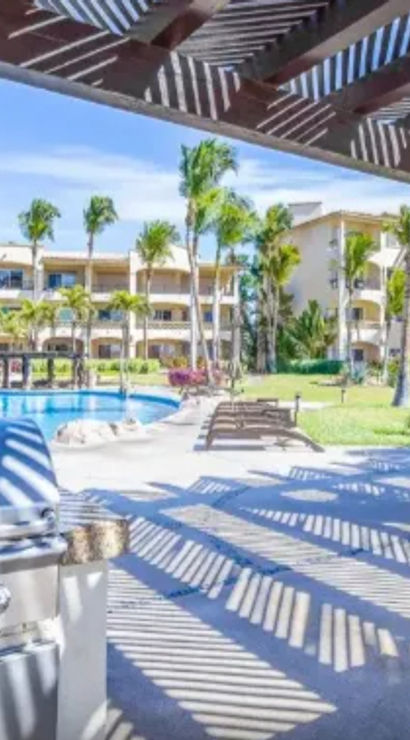 Sunlit beachfront resort patio with striped pergola shadows, stainless-steel grill, poolside lounge chairs, tropical palm trees and ocean view