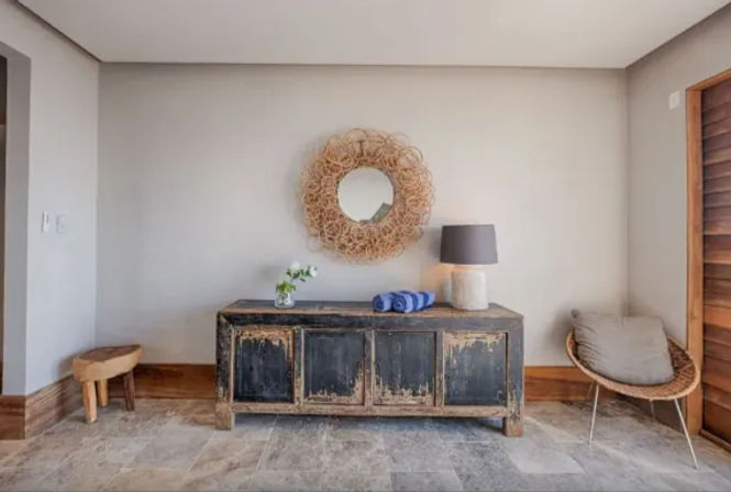 Modern rustic foyer with distressed black console table, round woven mirror, gray lamp, blue rolled towels and wicker chair on stone tile floor.