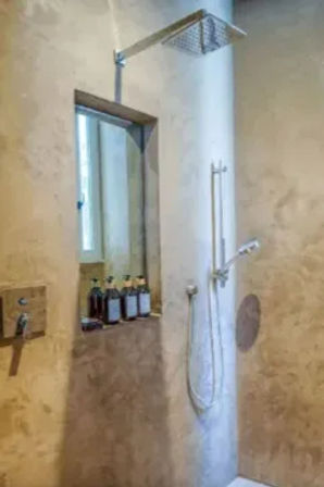 Beige plaster shower with ceiling-mounted square rain showerhead, wall-mounted handheld spray, and a recessed niche holding four amber pump bottles