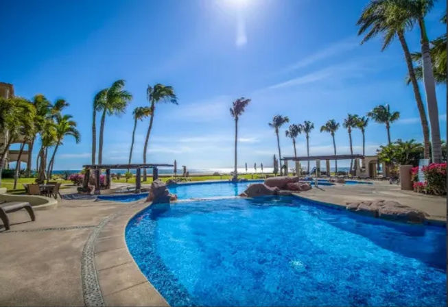 Crystal-blue oceanfront resort pool with rock features and cabanas, palm trees swaying under a sunny sky overlooking the beach.